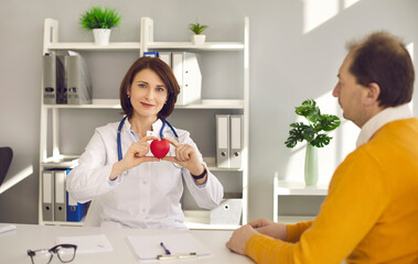 Happy doctor holding red heart and looking at camera sitting at office desk with senior patient. Cardiovascular disease treatment, heart attack preventive medical care, maintaining good health concept