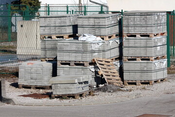 Grey stone tiles and concrete curbs stacked on top of wooden pallets at local construction site surrounded with paved road and metal fence in background on warm sunny winter day