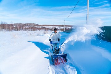 A man in a white jacket removes snow from a rural road with a blue snowblower in winter after a snowfall - 416492823