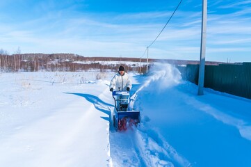 A man in a white jacket removes snow from a rural road with a blue snowblower in winter after a snowfall - 416492826