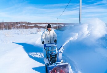 A man in a white jacket removes snow from a rural road with a blue snowblower in winter after a snowfall