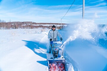 A man in a white jacket removes snow from a rural road with a blue snowblower in winter after a snowfall - 416492849