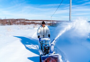 A man in a white jacket removes snow from a rural road with a blue snowblower in winter after a...