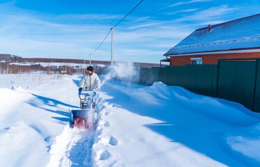 A man in a white jacket removes snow from a rural road with a blue snowblower in winter after a snowfall - 416492855