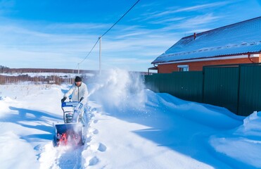 A man in a white jacket removes snow from a rural road with a blue snowblower in winter after a snowfall - 416492862
