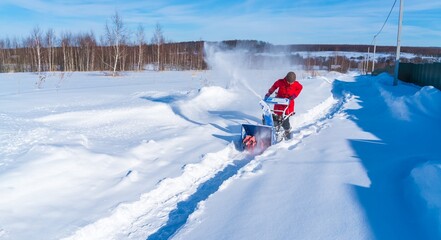 A woman in a red jacket removes snow from a rural road with a snowblower in winter after a snowfall - 416492863