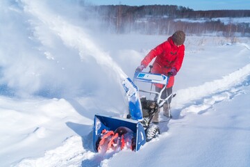 A woman in a red jacket removes snow from a rural road with a snowblower in winter after a snowfall - 416492879