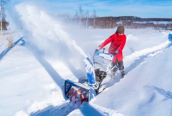 A woman in a red jacket removes snow from a rural road with a snowblower in winter after a snowfall