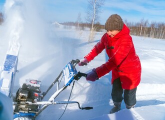 A woman in a red jacket removes snow from a rural road with a snowblower in winter after a snowfall - 416492881