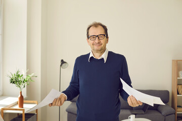 Smiling mature man in eyeglasses standing in his workplace and holding paper documents in both...