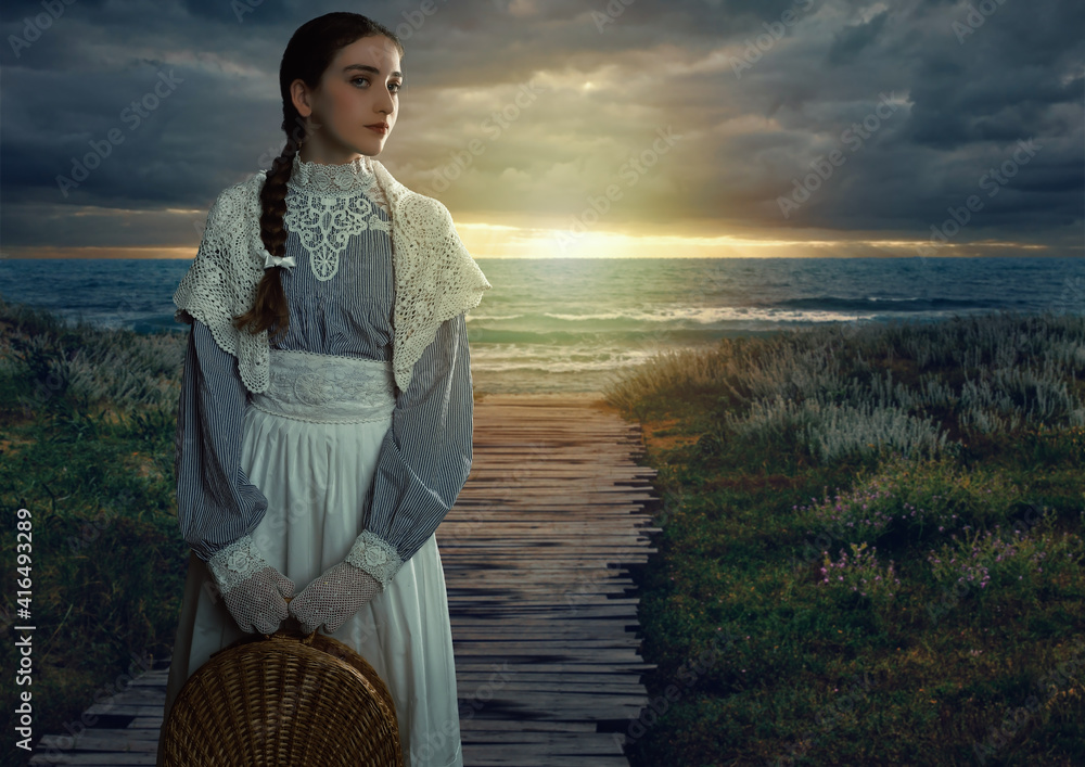 Wall mural Victorian girl with a hood and a wicker basket in her hands dressed in a white dress and blue striped blouse stands on the seashore at sunset.