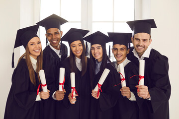Successful graduation from college or university. Group of young smiling multiethnic university graduates in black mantles standing with diplomas in hands and looking at camera in university classroom