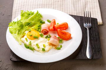 Scrambled eggs with bacon garnished with tomato and green onions and salad on a gray concrete background on a wooden stand next to a fork.