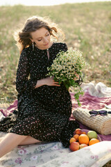 An attractive young pregnant woman in a light dress is resting on a picnic in nature. Expectant mother waiting for childbirth. The beginning of the third trimester of pregnancy