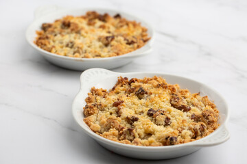 Two plates of delicious apple crumble pie isolated on marble background. Close-up of double crumble pies with fruits, nuts and cinnamon in white bowls. Dessert menu concept.
