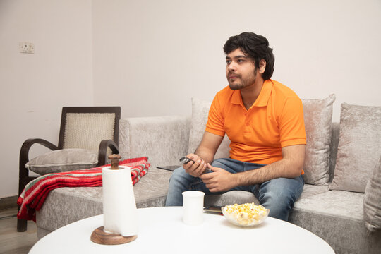 Asian Indian Man Gaming, Streaming And Watching TV Alone In Living Room On A Grey Sofa And White Table