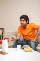 Asian Indian man gaming, streaming and watching TV alone in living room on a grey sofa and white table