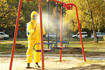 Person in hazmat suit with disinfectant sprayer cleaning children's playground. Surface treatment during coronavirus pandemic
