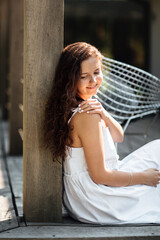 Fototapeta premium Portrait of a carefree young lady in white dress enjoying free time during summer vacation sitting on terrace in exterior, cute woman feeling so happy smiling at home.