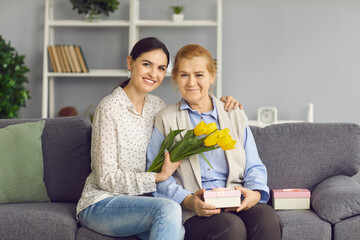 Portrait of daughter and old mother sitting on sofa with gifts and flowers, smiling and looking at camera. Young and senior women enjoying Women's Day. Happy beautiful grandma gets birthday presents