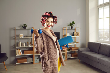 Cheerful young woman wants to keep fit and enjoys fitness exercise at home. Happy smiling housewife in curlers and beauty face mask standing in living-room and holding workout dumbbells and sports mat