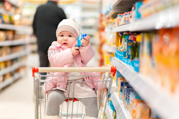 Shopping. A cute child is sitting in a shopping cart in the middle of a supermarket and holding a product. The concept of family shopping and parenting © _KUBE_