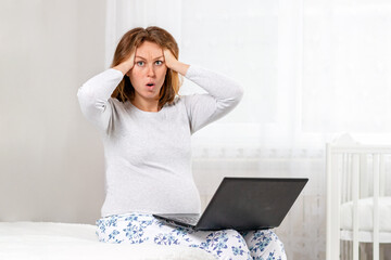 A pregnant woman is sitting on a bed with a laptop on her lap, holding her head with her hands in shock. White room in the background. The concept of the Internet and social networks