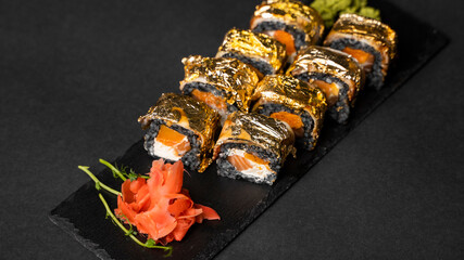 Custom sushi roll with nori, cuttlefish ink, fresh salmon, cream cheese, pepper pumpkin, eel, unagi sauce and edible gold leaf with red ginger, wasabi on a square plate on black table and background.