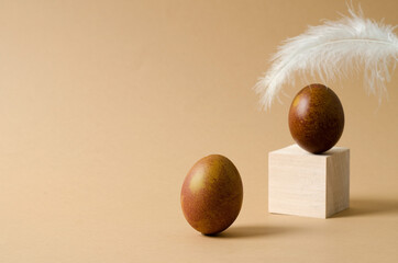 Minimalistic Easter background. A brown chicken egg stands on a beige background, an egg and a feather are out of focus in the background. Copy space