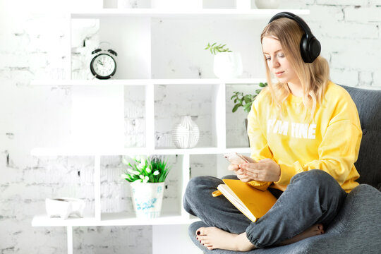 technology, online education, video blog concept. Girl in earphones in yellow blouse sit relax on grey couch use smartphone and note to listen music, courses, wish list. White brick wall background