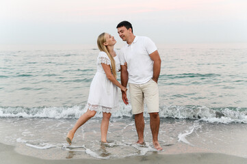 beautiful girl with white hair kisses her husband on the cheek on the beach near the ocean at sunset. romantic walk. vacation of a young family. travel to the sea.