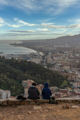 A couple from the top of the hill contemplating the beautiful views of the city of Malaga with the mountains and the cloudy sky in the background.