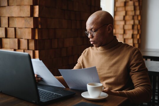 Portrait of smiling African American man in glasses sit at desk in office working on laptop, happy biracial male worker look at camera posing, busy using modern computer gadget at workplace