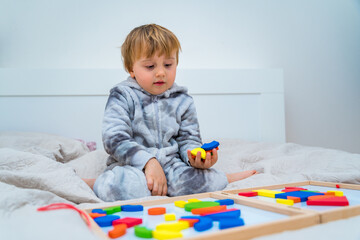 Little toddler boy plays on the bed in a wooden magnetic constructor, preschool child learning at home to make mosaic. Early development. Playing with chikldren during coronavirus outbreak.