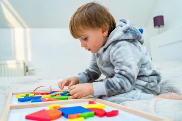 Little toddler boy plays on the bed in a wooden magnetic constructor, preschool child learning at home to make mosaic. Early development. Playing with chikldren during coronavirus outbreak.