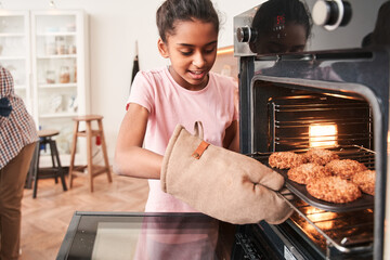 Girl taking tray with cookies out of the oven while baking at the kitchen