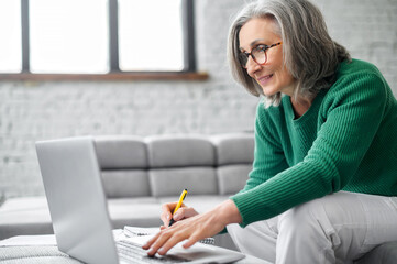 Charming senior online teacher conducts webinar, online classes, mature modern elderly woman sitting with a laptop at home, studying, taking a notes. An elderly modern business lady works remotely