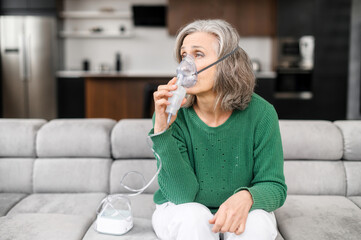 Sick senior woman has a viral infection or flu, labored breathing, asthma. An elderly female is using inhaler, nebulizer for self treatment sitting alone on the sofa at home - obrazy, fototapety, plakaty