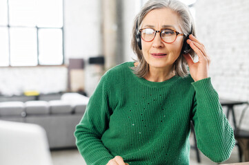 Concentrated senior woman wearing wireless headset takes on call, a mature female support employee...
