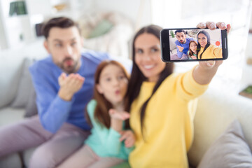 Photo of young cheerful family mom make selfie smartphone dad daughter send air kiss happy positive smile sit sofa home