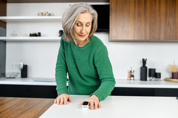 Smiling senior woman using pulse oximeter at home, measuring oxygen saturation by herself, pulse...
