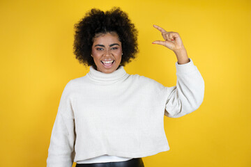 African american woman wearing casual sweater over yellow background smiling and confident gesturing with hand doing small size sign with fingers . Measure concept.