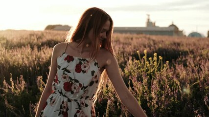 A young red-haired girl happily walking in slow motion through green field. Beautiful carefree woman enjoying nature and sunlight at incredible colorful sunset.