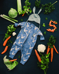 Adorable little child boy in pajamas with a rabbit and a hat with ears on a background of green vegetables. The kid begins to eat adult food. Easter style. 