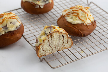 Close-up of delicious cinnabons on a cooling tray on white marble background. One is cut in half. Cinnamon rolled buns with white sticky glaze, almond flakes and caramel on top. Dessert buns pastry.
