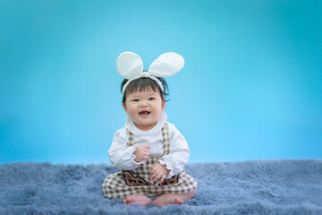 Baby in Easter bunny hairband. Asian happy baby smiling and sitting on carpet on blue background. Cute 6 months baby start sitting with copy space as Easter concept, baby or kid department in hospital