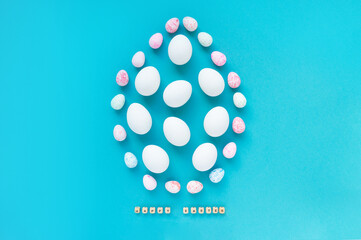 White chicken eggs and small colored eggs are laid out in the form of a large egg in the center on a blue background. Easter creative design, greeting card. The inscription Happy Easter. Top view