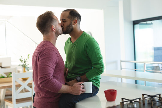 Multi ethnic gay male couple sitting in kitchen drinking coffee and kissing at home