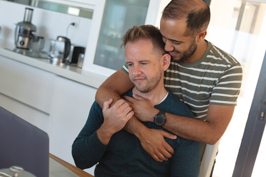 Multi ethnic gay male couple eating breakfast embracing and using laptop at home