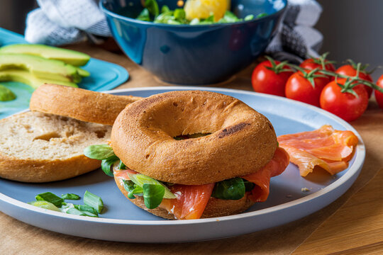 Bagel sandwiches in the making. Gluten free toasted bagel with smoked salmon, cream cheese and lettuce. Avocado, tomatoes and cucumber in background. Homemade Healthy breakfast with fun vibrant colors - Powered by Adobe